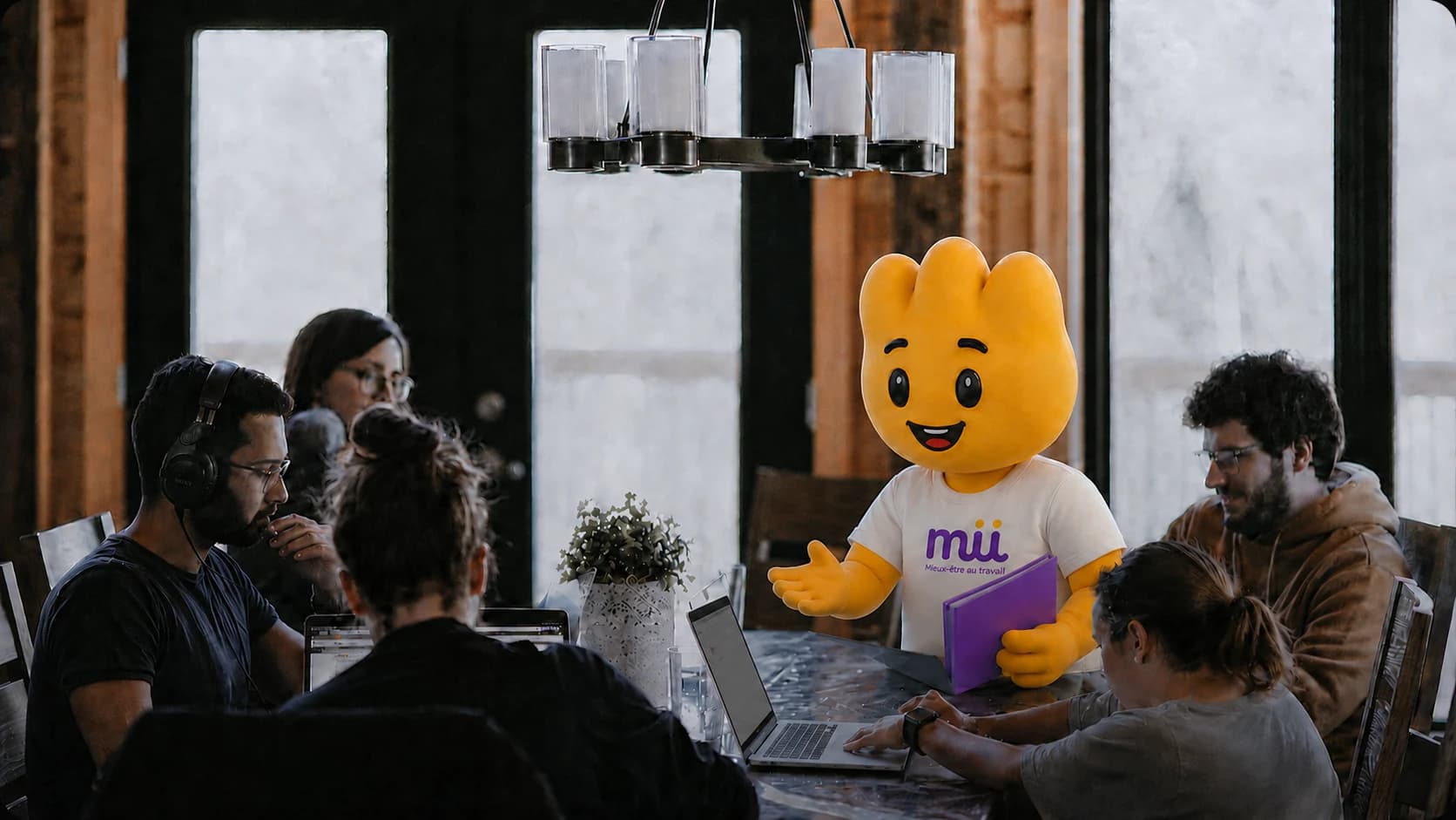 La mascotte Mü échange avec une équipe réunie autour d’une table de travail.